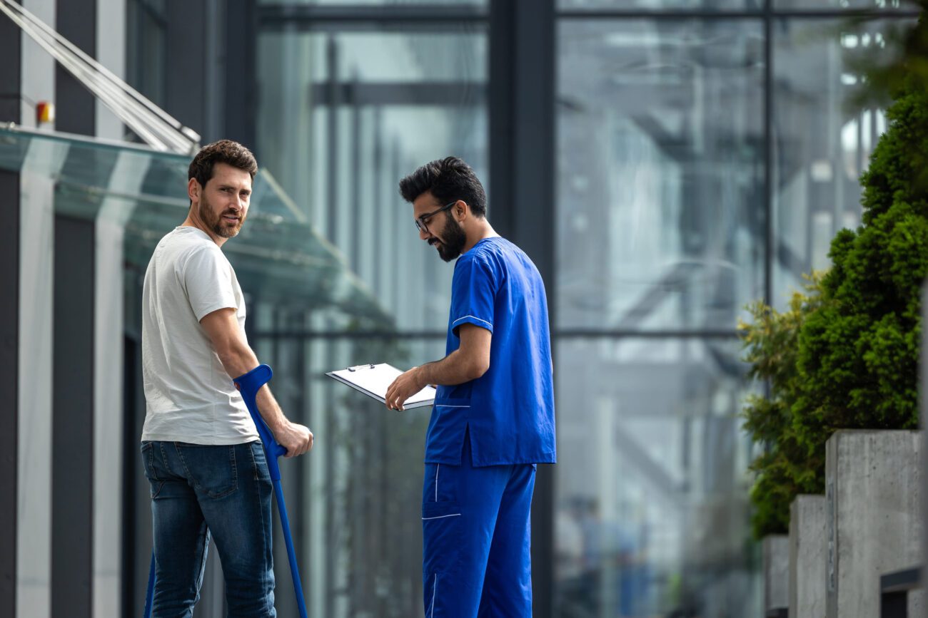 Man with a blue forearm crutch speaks with a clinician in blue scrubs who holds a clipboard outside a glass-walled building.&nbsp;Hispamer Noticias