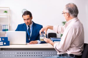 Two men in an office discuss savings; one places coins into a jar labeled SAVINGS during a meeting at a desk with a laptop.&nbsp;Hispamer Noticias