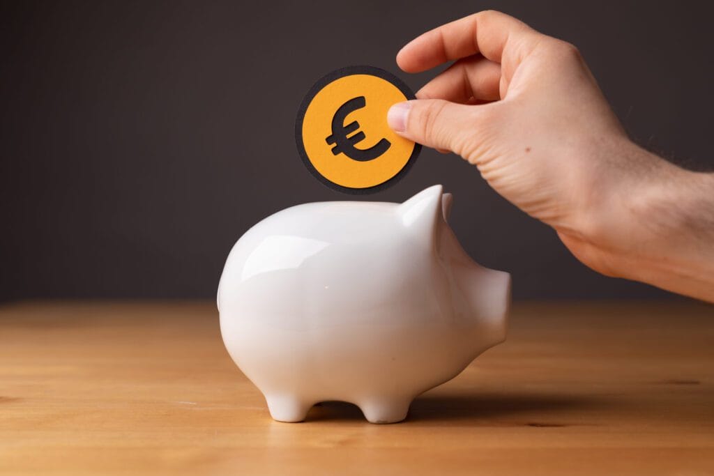 Hand placing a euro coin into a white piggy bank on a wooden surface, symbolizing saving money.&nbsp;Hispamer Noticias