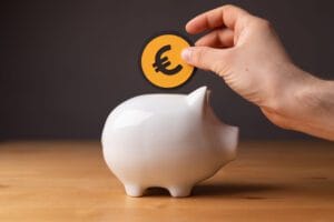 Hand placing a euro coin into a white piggy bank on a wooden surface, symbolizing saving money.&nbsp;Hispamer Noticias