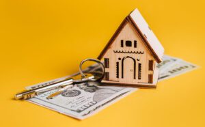 Wooden house model resting on dollar bills with keys, symbolizing real estate or mortgage costs on a yellow background.&nbsp;Hispamer Noticias