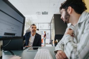 Two coworkers discuss at a desk in a bright office; one man in a suit talks to a colleague leaning in nearby computer setup.&nbsp;Hispamer Noticias