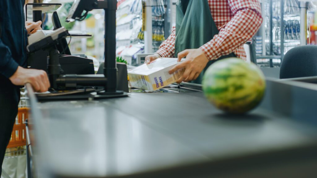 Store checkout: cashier scans a box as a customer places it on the conveyor; a watermelon sits on the belt in the foreground.&nbsp;Hispamer Noticias