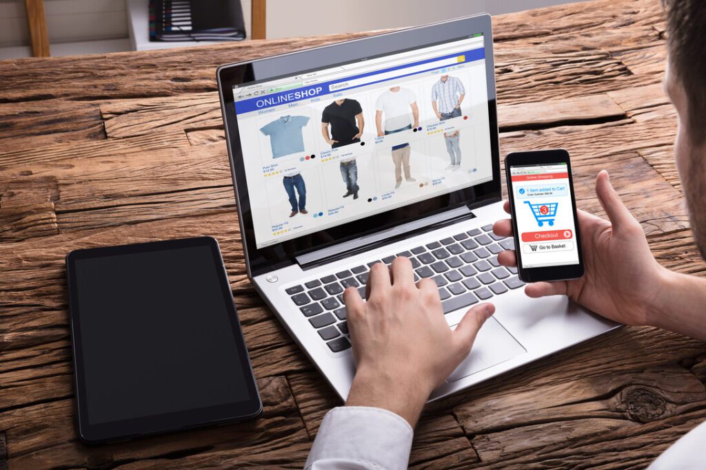 Person shops online on a laptop and smartphone, browsing clothing items on an e-commerce site.&nbsp;Hispamer Noticias