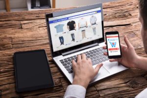 compras online importacion - Hispamer Noticias Person shops online on a laptop and smartphone, browsing clothing items on an e-commerce site. Hispamer Noticias