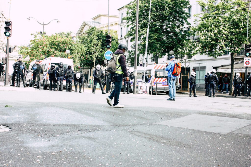 Line of riot police in protective gear blocking a city street beside plastic barricades, with a passerby carrying a backpack nearby.&nbsp;Hispamer Noticias